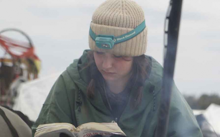 a student wearing winter clothing reads from a book while sitting amongst a snowy landscape